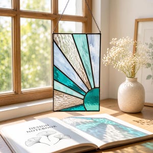 Op de afbeelding: Een rechthoekige glas-in-lood zonnevanger met een zonnestraalpatroon in tinten van talingblauw, lichtblauw en helder getextureerd glas. Het object is omlijst met donker metaal en hangt aan een dunne ketting, met een geometrische indeling.