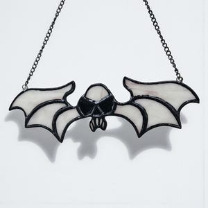 May include: A stained glass bat-shaped suncatcher featuring a skull design. The bat has white wings and a black outline, with a black skull in the centre. It hangs from a black chain, ideal for Halloween or gothic decor.