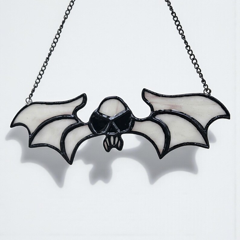 May include: A stained glass bat-shaped suncatcher featuring a skull design. The bat has white wings and a black outline, with a black skull in the centre. It hangs from a black chain, ideal for Halloween or gothic decor.