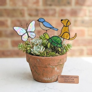 Puede incluir: Una maceta de terracota llena de suculentas y figuras decorativas de vidrio de una mariposa, un pájaro, una tortuga y un perro. La mariposa tiene alas iridiscentes. La etiqueta dice "Handmade Artisan Collection - Suzy Glass."