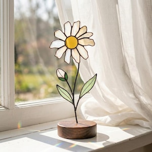 Puede incluir: Una escultura de margarita de vidriera con centro amarillo y pétalos blancos. La flor se asienta sobre un fino tallo negro con hojas verdes y un capullo. La base es un bloque de madera redondo y oscuro. La escultura se exhibe cerca de una ventana.