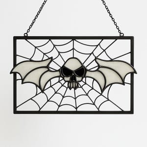 May include: A black framed Halloween decoration featuring a bat-skull design with a spiderweb background. The bat-skull has glowing white wings and a skull face with black eyes. The decoration hangs from a chain.