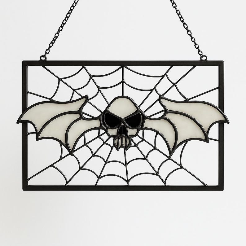May include: A black framed Halloween decoration featuring a bat-skull design with a spiderweb background. The bat-skull has glowing white wings and a skull face with black eyes. The decoration hangs from a chain.