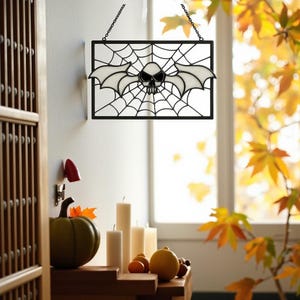 May include: A rectangular Halloween stained glass window hanging from a chain. The design features a skull with bat wings in the center of a spider web. The window is black with white and clear glass. A green pumpkin and candles are on a wooden surface.