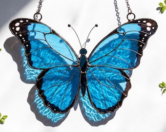 May include: A stained glass butterfly suncatcher featuring vibrant blue wings with black borders and white spots. The wings are framed in dark metal solder lines, and the piece includes two thin metal antennae and a hanging chain at the top.