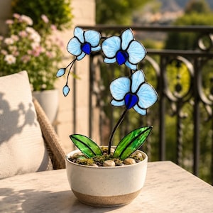 Könnte beinhalten: Eine dekorative Glasorchidee in einem weißen Keramiktopf. Die Orchidee hat hellblaue Blütenblätter mit dunkelblauen Zentren, grüne Blätter und einen schwarzen Stiel. Der Topf ist mit Moos und kleinen Steinen gefüllt.