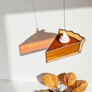 May include: A stained glass pie slice with a dollop of white whipped cream. The pie has a golden crust and a filling that transitions from orange to amber. The piece is suspended by a thin string, casting a shadow on the white background.