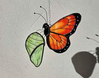 May include: Stained glass butterfly suncatcher with orange and black wings, and a green chrysalis. The butterfly is suspended by a silver chain. The suncatcher is designed to hang and catch the light.