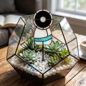 May include: Geometric glass terrarium with succulents and white pebbles. A decorative stained glass figure with black, white, and turquoise accents is placed in the center. The terrarium sits on a wooden table, with a coffee cup and a window in the background.