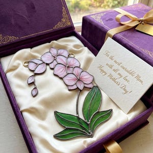May include: A stained glass orchid in a purple velvet gift box. The orchid features pink petals and green leaves. A card reads, "For the most incredible Mom, with all my love. Happy Mother's Day!"