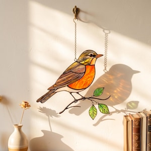 May include: A stained glass robin suncatcher, with orange, brown, and yellow plumage, perched on a branch with green leaves. It hangs from a chain and hook. The decorative bird casts a shadow on the wall, a charming piece for home decor.