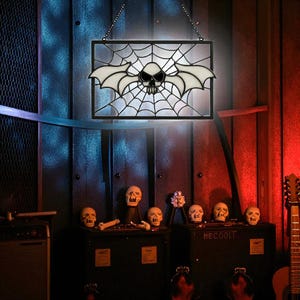 May include: A Halloween stained glass window with a black frame, depicting a skull with bat wings set against a spiderweb design. The window hangs from a chain. Decorative skulls and a guitar are also visible.