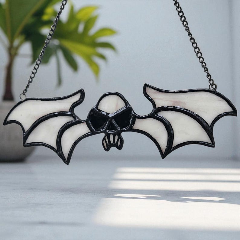 May include: A stained glass bat ornament featuring a skull design. The bat has white wings with black edging and a black skull face. It hangs from a black chain. A decorative item, possibly for Halloween or gothic home decor.