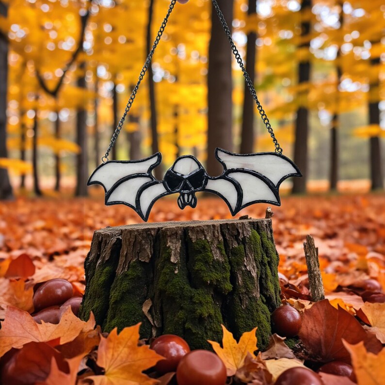 May include: A stained glass bat decoration in black and white, featuring a skull design, suspended by a chain. The bat is set against a backdrop of autumn leaves and trees, resting on a tree stump.