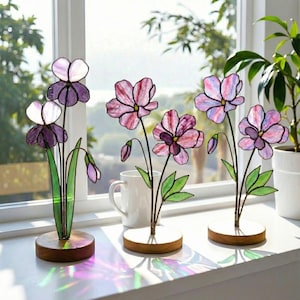 May include: Stained glass flower sculptures in shades of purple, pink, and green, with brown wooden bases. The flowers are displayed on a windowsill, with a white mug and a potted plant in the background. The light shines through the glass, creating colorful reflections.