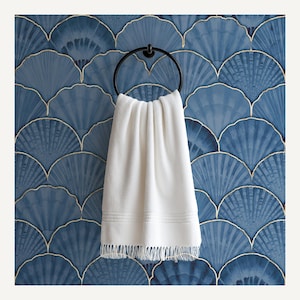 Op de afbeelding: Een witte handdoek met franjes hangt aan een zwarte metalen ring. De handdoek bevindt zich tegen een blauwe tegelwand met een waaiervormig ontwerp en gouden randen. Moderne badkamerdecoratie.