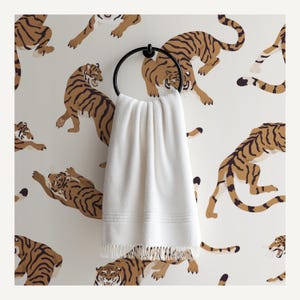 May include: A white towel with a fringed edge hangs from a black circular towel ring. The background features a repeating pattern of brown and black tigers on a cream background. The towel has a textured design.