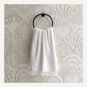 Op de afbeelding: Een witte badhanddoek met franjes hangt aan een zwarte metalen handdoekring. De handdoek heeft een textuuroppervlak en decoratieve stiksels. De achtergrond heeft een waaierpatroon behang in beige en wit.
