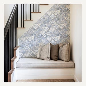 Pode incluir: Um banco aconchegante sob uma escada com papel de parede com padr&atilde;o de conchas em tons de azul e branco. O banco tem uma almofada cinza claro e tr&ecirc;s almofadas decorativas em tons neutros. Corrim&atilde;o preto e escada de madeira.