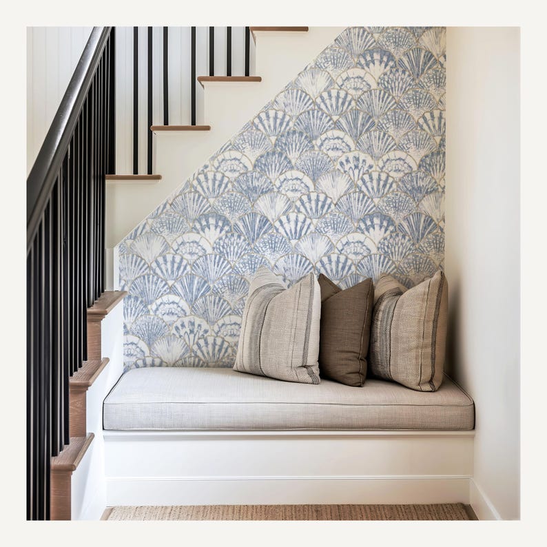 Pode incluir: Um banco aconchegante sob uma escada com papel de parede com padr&atilde;o de conchas em tons de azul e branco. O banco tem uma almofada cinza claro e tr&ecirc;s almofadas decorativas em tons neutros. Corrim&atilde;o preto e escada de madeira.