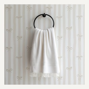 May include: A white towel with fringe hangs from a black metal ring. The towel is displayed against a striped wallpaper background with bow patterns in a neutral color palette. The towel has a textured appearance.
