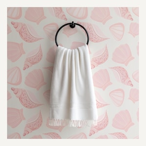 May include: A white towel with fringe hangs from a black metal ring. The towel is displayed against a wall covered in a repeating pattern of pink seashells on a white background. The towel has a textured appearance.