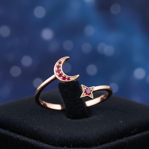 Puede incluir: Un anillo de oro rosa con un diseño de luna creciente y estrella, ambos adornados con pequeñas gemas rojas. El diseño abierto permite un ajuste ajustable. El anillo se muestra sobre un fondo oscuro.