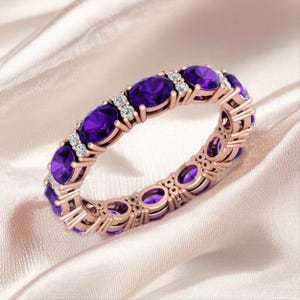 May include: Rose gold ring featuring oval-cut purple gemstones and small white accent stones. The ring is set on a white satin surface. The gemstones are evenly spaced around the band, creating a continuous design.