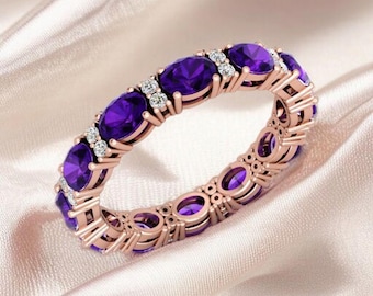 May include: Rose gold ring with oval purple gemstones and small white accent stones. The ring is set on a white satin surface. The gemstones are evenly spaced around the band, creating a continuous design.