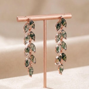 May include: Rose gold earrings with a leaf design. Each earring has multiple leaf-shaped pendants with green and white marbling. The earrings are displayed on a rose gold stand.
