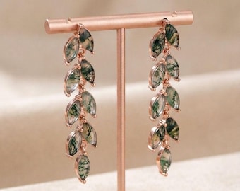 May include: Rose gold earrings with a leaf design. Each earring has multiple leaf-shaped pendants with green and white marbling. The earrings are displayed on a rose gold stand.