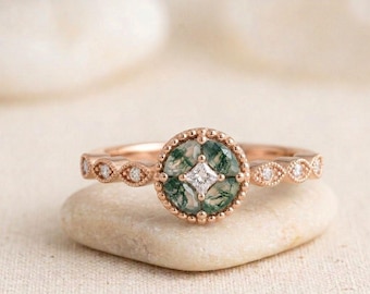 May include: A rose gold ring featuring a circular moss agate centre stone with a small square-cut diamond at its core. The band is detailed with a beaded edge and small round diamonds set within marquise-shaped metal frames along the shank.