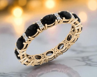 May include: A gold ring set with oval black gemstones and small, clear accent stones. The ring's band has an openwork design. The background is blurred with warm tones. The ring is a piece of jewellery.