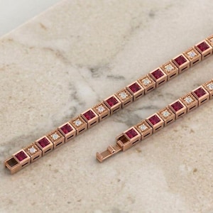May include: A rose gold tennis bracelet featuring alternating square-cut deep red rubies and small round white diamonds set in individual metal frames. The bracelet includes a secure box clasp mechanism with a fold-over safety latch.