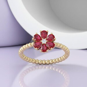 May include: A gold ring featuring a flower design with red gemstones and a central diamond. The ring's band has a textured, beaded appearance. The flower petals are a deep red, and the central diamond sparkles.