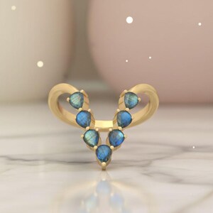 May include: A gold-colored ring with a V-shaped design. The ring is adorned with seven pear-shaped, blue gemstones. The gemstones are arranged in a symmetrical pattern, creating a unique and elegant look. The ring is set against a soft, blurred background.