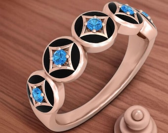 May include: Rose gold ring featuring a series of black circular accents with a square, white border and a central, round, light blue gemstone. A small, rose gold, tiered object sits to the side.