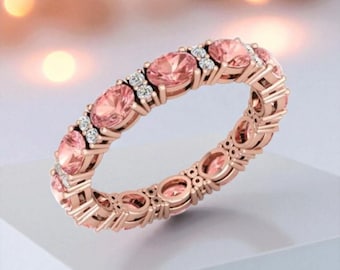 May include: A rose gold eternity ring with oval-cut pink gemstones and small white accent stones. The ring's band is also rose gold, creating a cohesive design. The ring is set against a soft, blurred background.