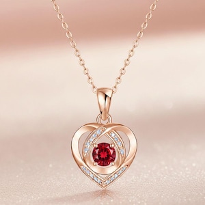 Puede incluir: Un collar de oro rosa con un colgante en forma de corazón. El colgante presenta una gema roja redonda central, rodeada por un diseño de doble corazón y pequeñas gemas claras. La cadena es delicada y combina con el color del colgante.