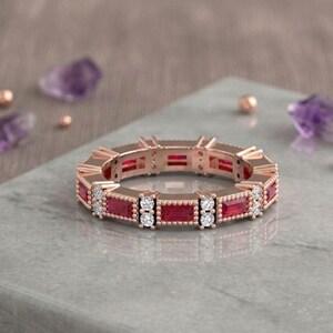 Peut inclure: Bague en or rose ornée de pierres rouges rectangulaires et de petits diamants blancs. La bague est posée sur une surface grise avec des cristaux violets et de petites perles dorées en arrière-plan. Le design de la bague est symétrique.