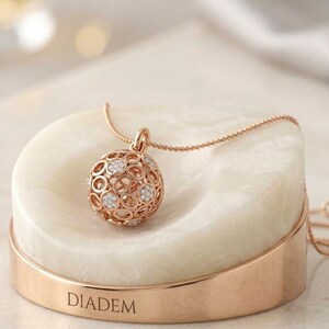 May include: A rose gold necklace featuring a spherical pendant with an openwork circular pattern and embedded clear stones. The pendant rests on a circular white stone base with a rose gold metal band engraved with the word DIADEM.