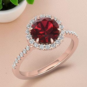 May include: Rose gold ring featuring a large, round, deep red gemstone surrounded by a halo of smaller, clear gemstones. The ring's band is also adorned with small, clear gemstones, creating a sparkling effect. The ring is set against a soft pink background.