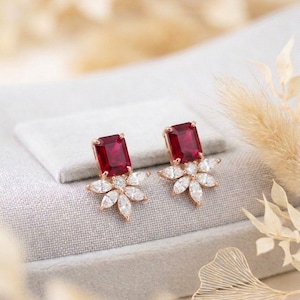 May include: A pair of rose gold earrings featuring a rectangular, deep red gemstone at the top. Below the red stone is a flower design with white, teardrop-shaped stones. The earrings are displayed on a gray surface.