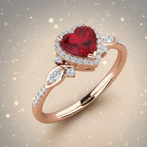 Peut inclure: Bague en or rose ornée d'une pierre rouge en forme de cœur, entourée de petits diamants transparents. D'autres diamants transparents ornent le corps et les côtés du cœur. La bague a une finition polie.