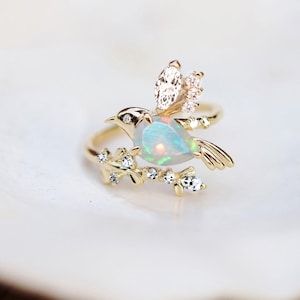 Könnte beinhalten: Ein goldener Ring mit einem Vogelmotiv, das einen Opalkörper und Diamantdetails aufweist. Der Vogel sitzt auf einem mit Diamanten geschmückten Ast. Der Ring hat eine polierte Oberfläche. Der Hintergrund ist weiß.