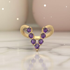 May include: A gold-colored ring featuring a V-shaped design adorned with seven purple gemstones. The ring has a smooth, polished finish and is set against a soft, blurred background.