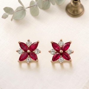 May include: A pair of flower-shaped stud earrings. Each earring features a cluster of red marquise-cut gemstones surrounded by smaller, clear round-cut stones. The earrings are set in a gold-colored metal and rest on a white surface.
