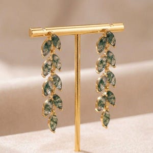 May include: Gold-coloured earrings with a leaf design. Each earring has multiple green and white marbled-effect leaf-shaped pendants hanging from a gold bar. The earrings are displayed on a gold stand against a neutral background.