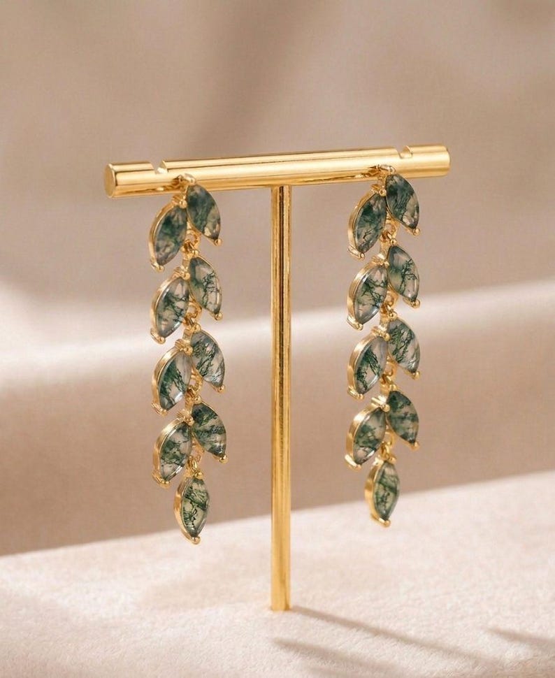 May include: Gold-coloured earrings with a leaf design. Each earring has multiple green and white marbled-effect leaf-shaped pendants hanging from a gold bar. The earrings are displayed on a gold stand against a neutral background.