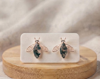 May include: A pair of bee-shaped stud earrings featuring rose gold-toned metal bodies and wings with a light pink enamel fill. The central abdomen of each bee is crafted from a marquise-cut moss agate gemstone with dark green inclusions.